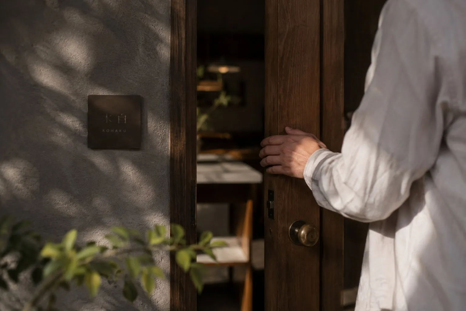 Une main pousse la porte d'un lieu, plaque gravée à l'entrée.
